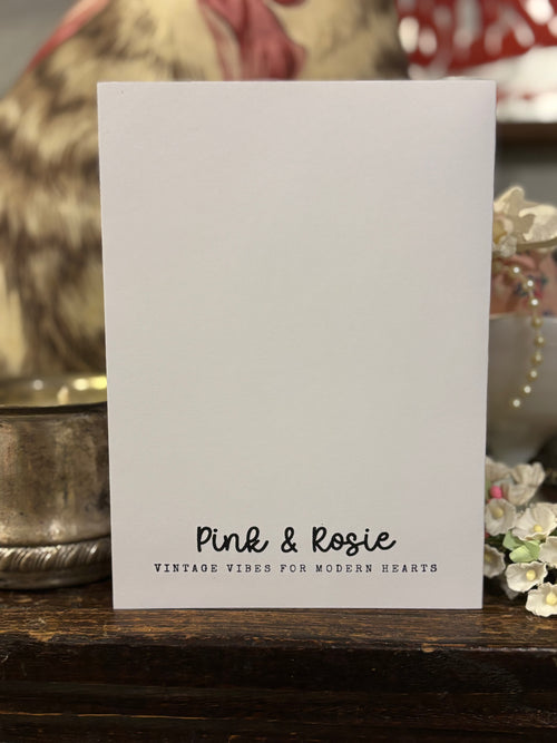 The back of the card is stamped in the color black that reads 'Pink & Rosie' and a tag line of "Vintage vibes for modern hearts" on a wooden surface with decorative items in the background