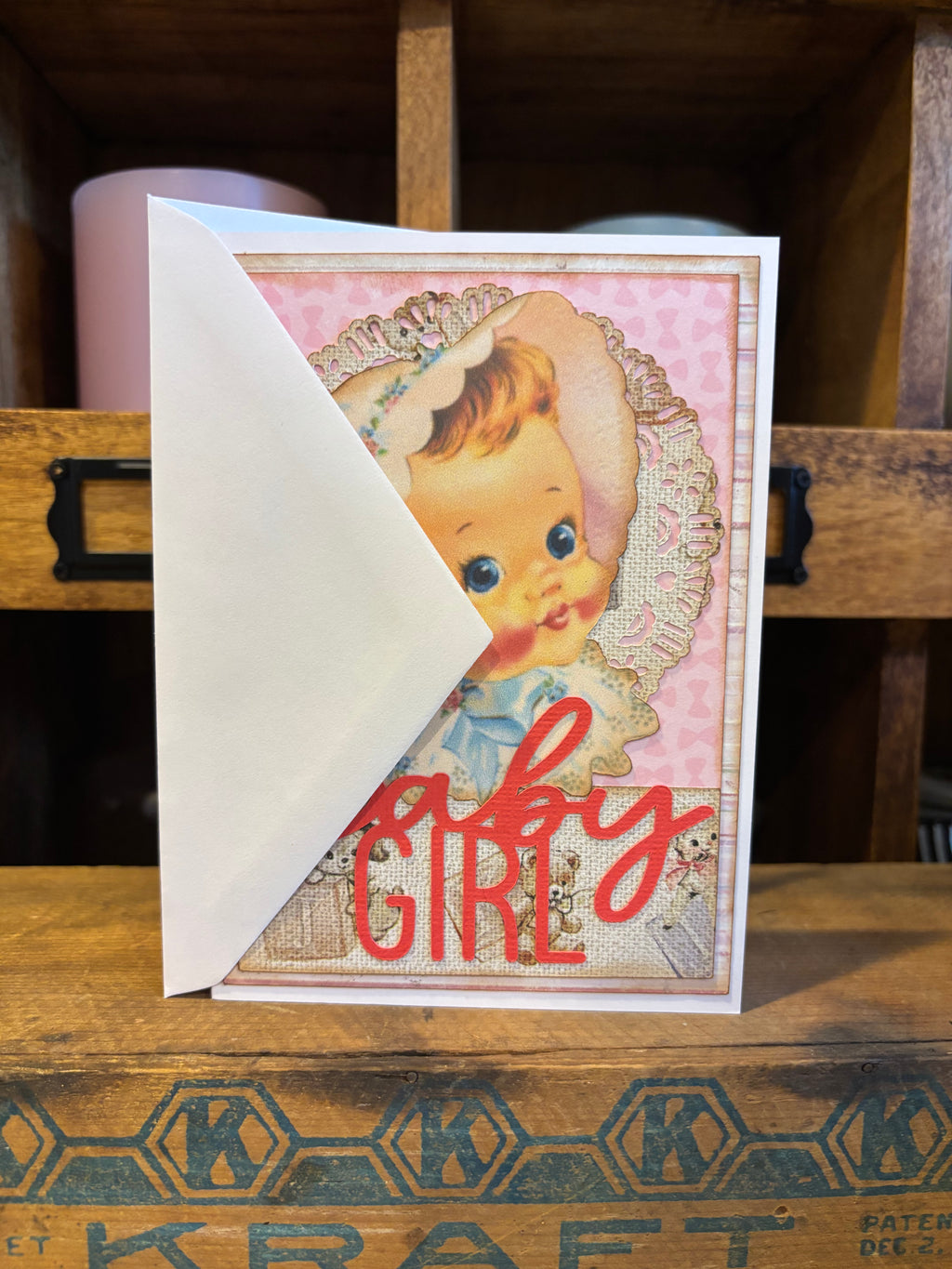 Vintage-style greeting card with a baby illustration and 'Girl' text on a wooden surface.