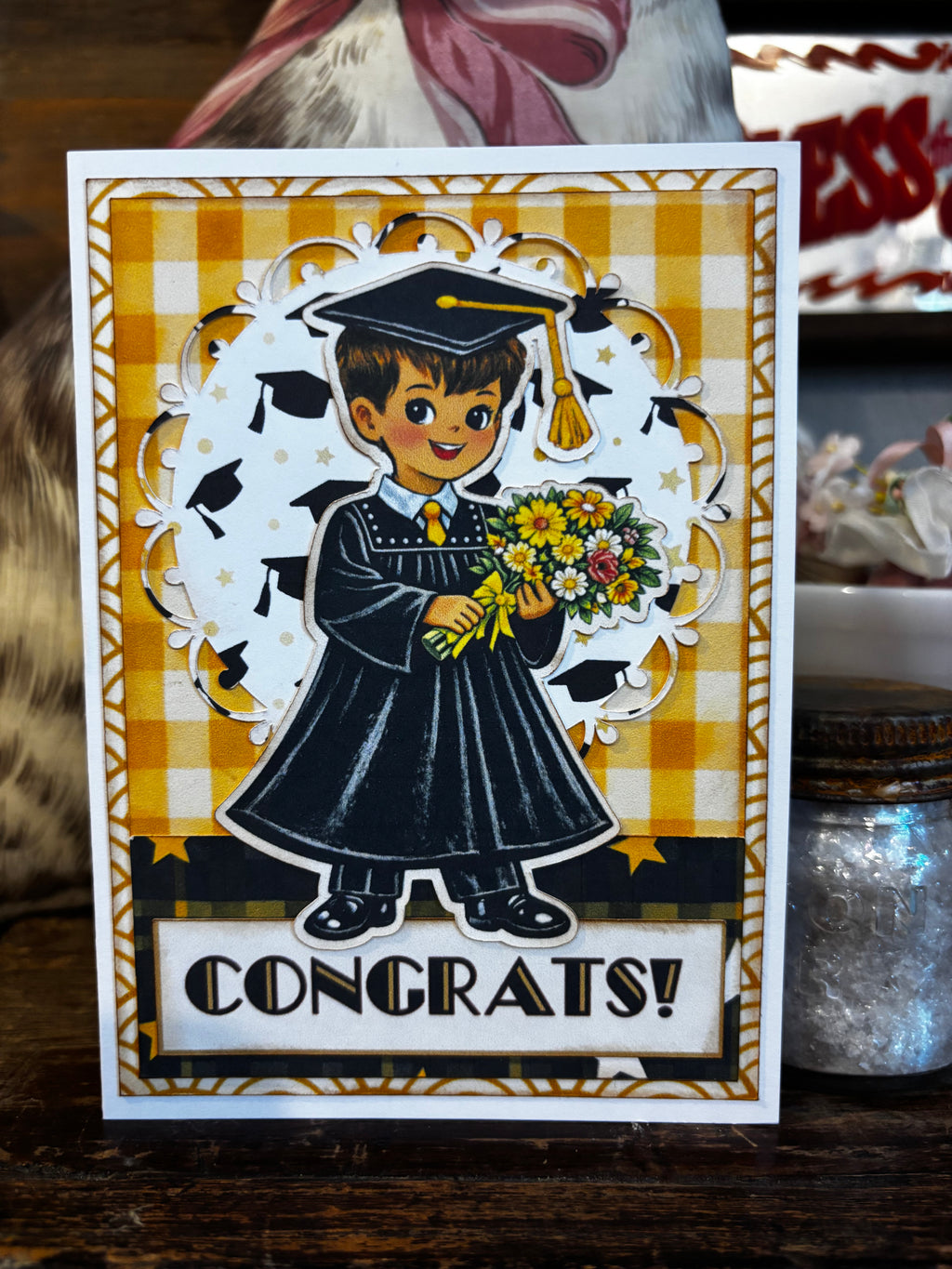 Graduation-themed card, boy/male holding flowers with decorative elements on a checkered background.