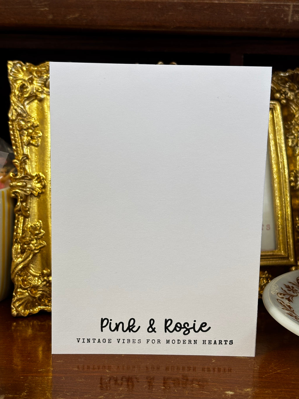 The back of the card is stamped in the color black that reads 'Pink & Rosie' and a tag line of "Vintage vibes for modern hearts" on a wooden surface with decorative items in the background