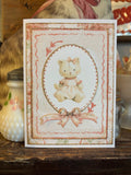 A card with a girl teddy bear in a pink-and-white dress, illustrated. There is gold glitter around the layers of paper on the card.