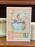 Vintage-style greeting card with a baby illustration and 'Girl' text, placed on a wooden surface with tea cups.